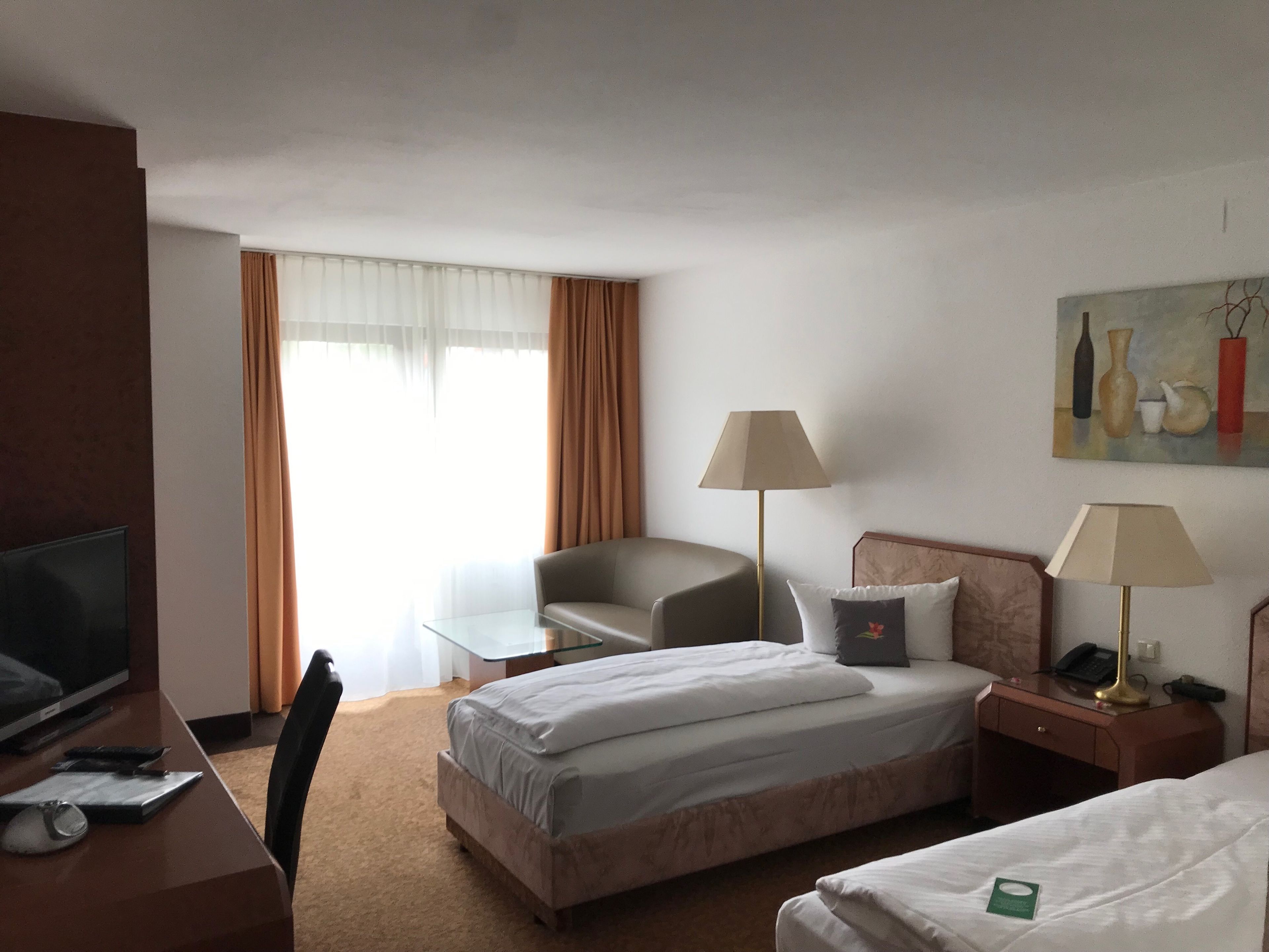 Ein Hotelzimmer mit einem Bett, einem Schreibtisch mit Fernseher, einem Sessel, einer Lampe und einem Bild an der Wand.