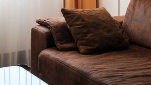 A brown leather sofa with two cushions, a floor lamp, and a glass coffee table in a room with sheer curtains and a white wall. - Hotel Flora Stuttgart - Möhringen (Stuttgart)