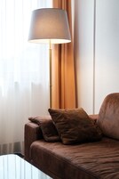 Un sofá de cuero marrón con dos cojines, una lámpara de pie y una mesa de centro de vidrio en una habitación con cortinas translúcidas y una pared blanca.
