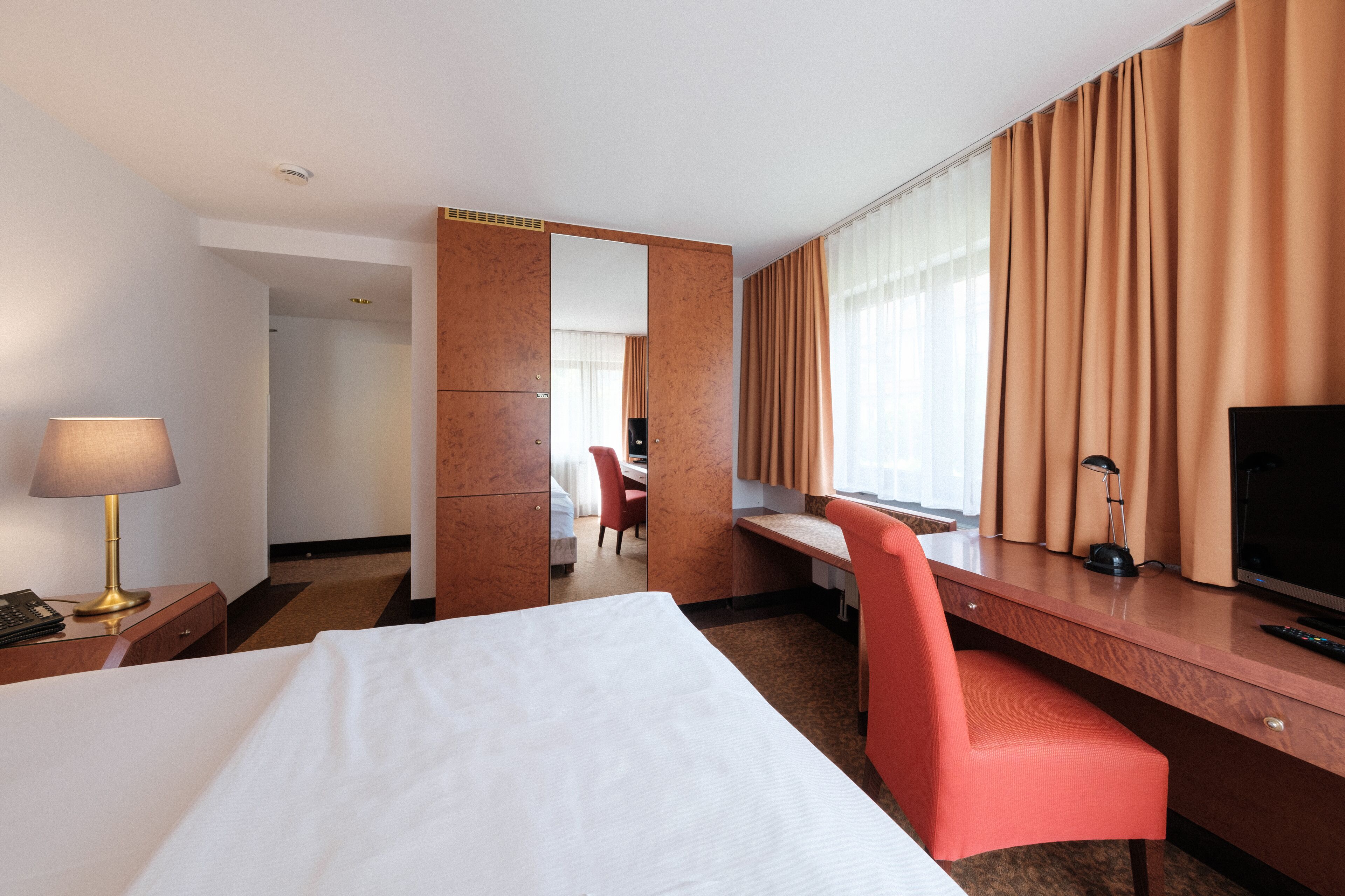 A hotel room with a bed, a desk, a chair, a TV, and a lamp.