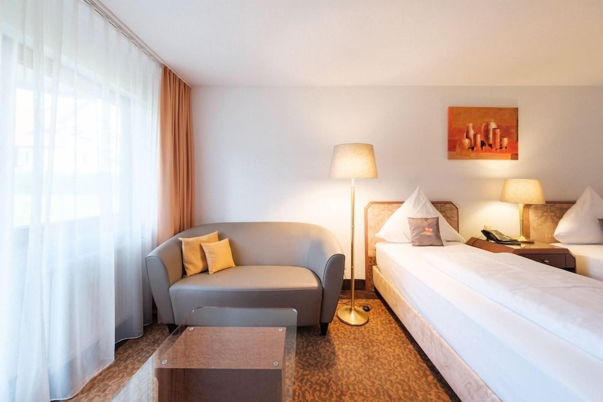 A hotel room with a sofa, a bed, a bedside table, a lamp, and a painting on the wall.