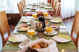 Una escena de desayuno con una mesa para dos, con platos, tazas, una cafetera y una cesta de pan.