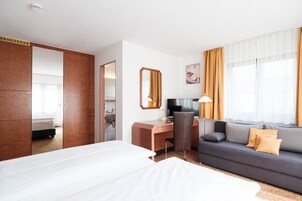 Habitación de hotel con cama, escritorio, silla, sofá y un amplio ventanal con cortinas.