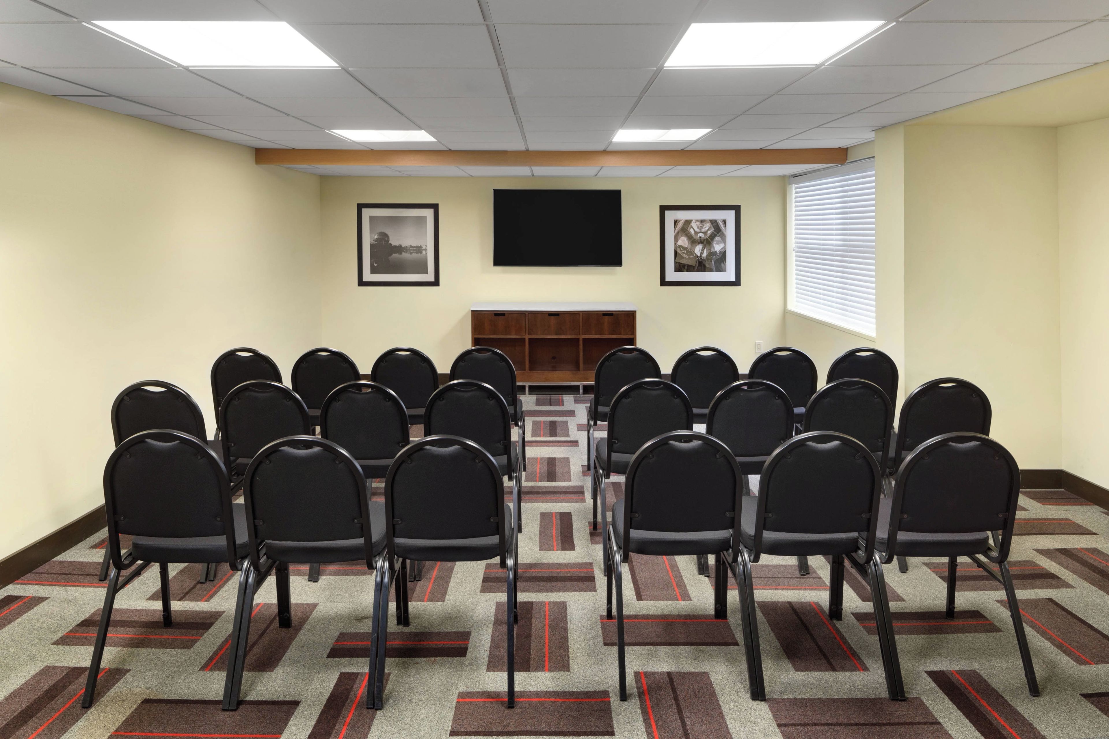 Une salle de conférence avec des rangées de chaises disposées face à une table centrale, un téléviseur à écran plat accroché au mur et des tableaux encadrés au-dessus de la table.