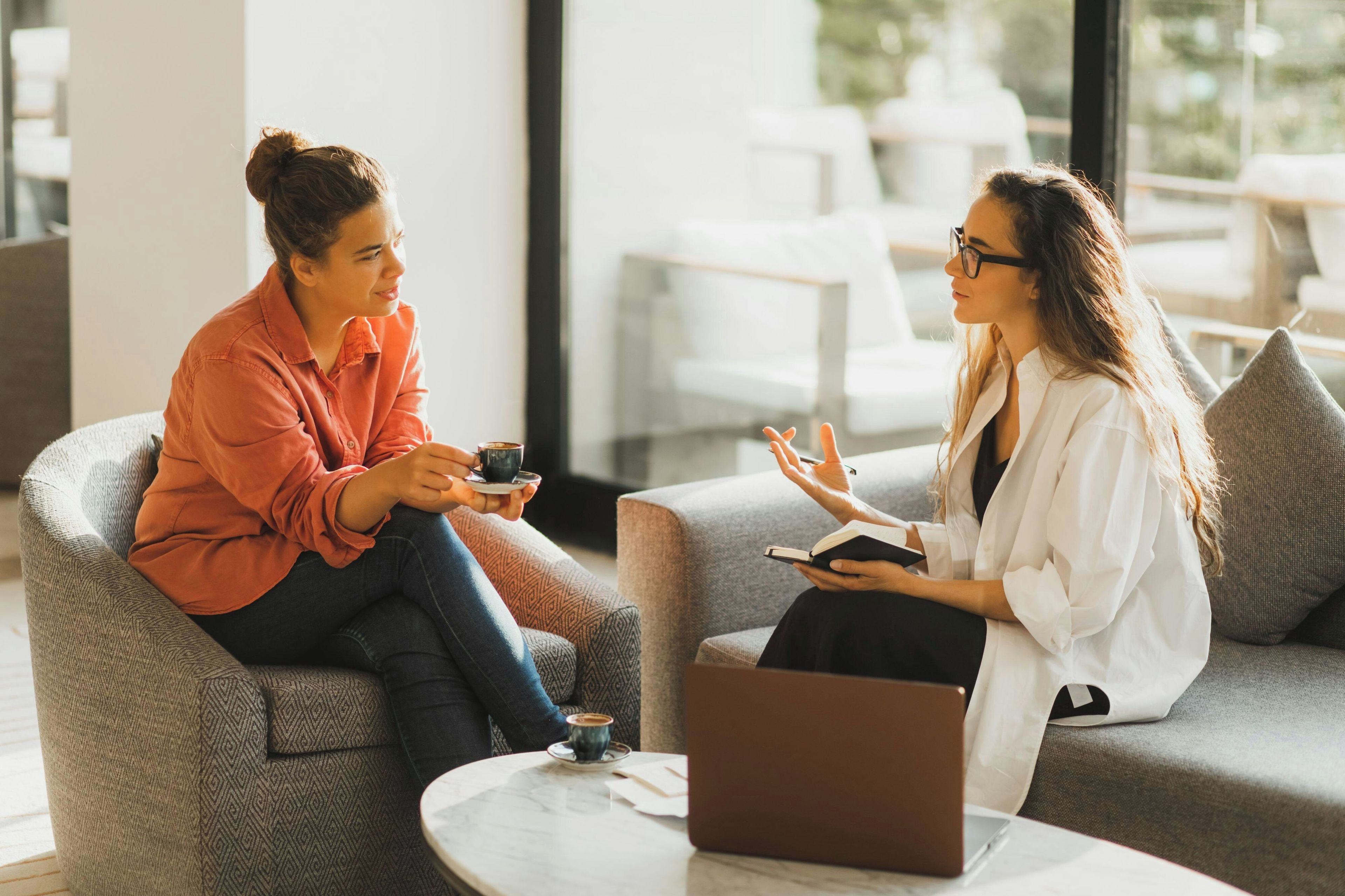 Dos personas sentadas en sillones estampados en un moderno salón con una mesa de mármol, una computadora portátil y una taza.