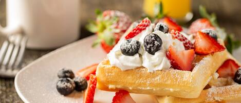 A plate of waffles topped with whipped cream, strawberries, and blueberries, with a cup of coffee and a glass of orange juice in the background.