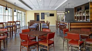 A spacious dining area with multiple tables and chairs, a bar area with shelves of bottles, and a large window providing natural light.