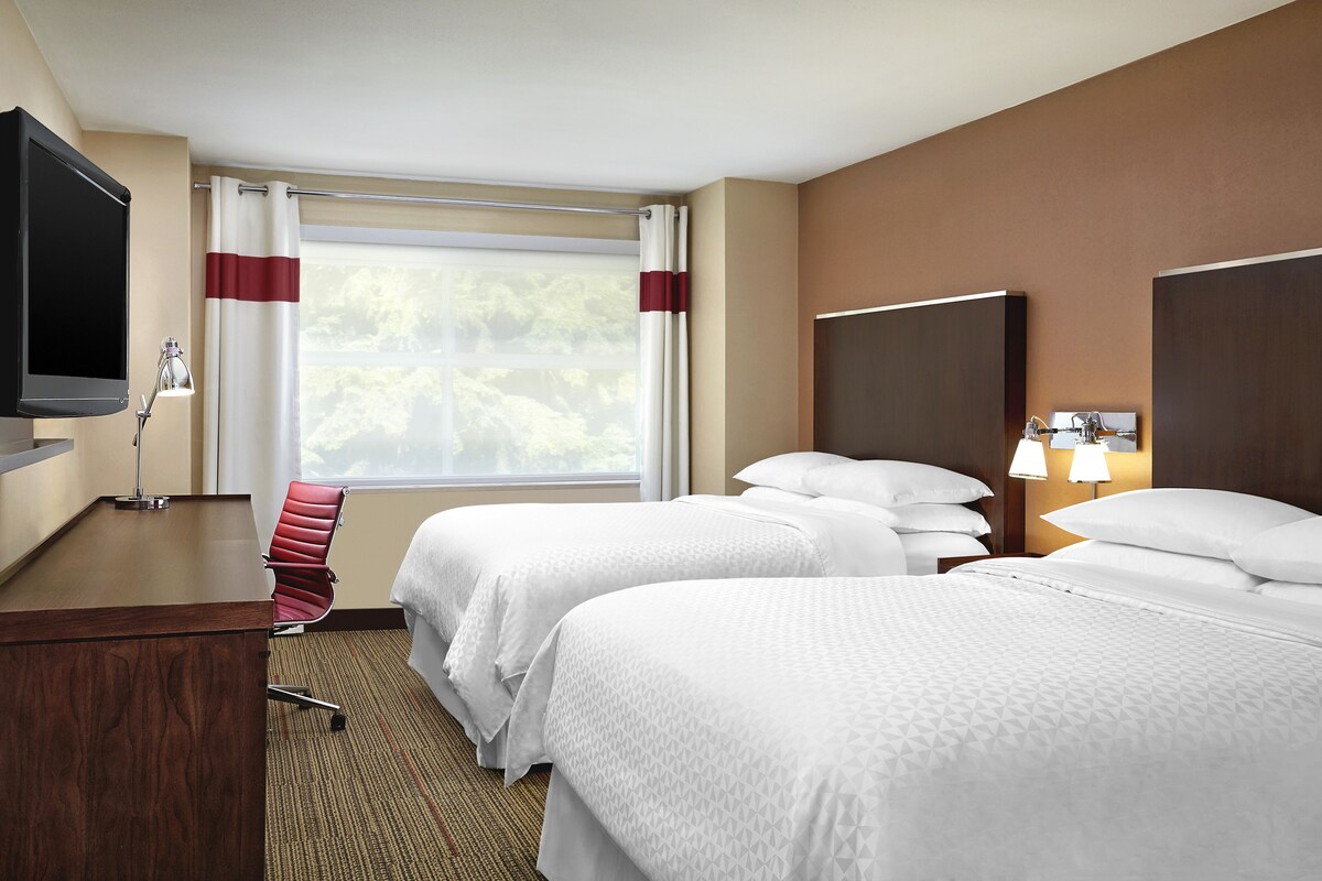 A hotel room with two beds, a desk, a TV, and a window with curtains.