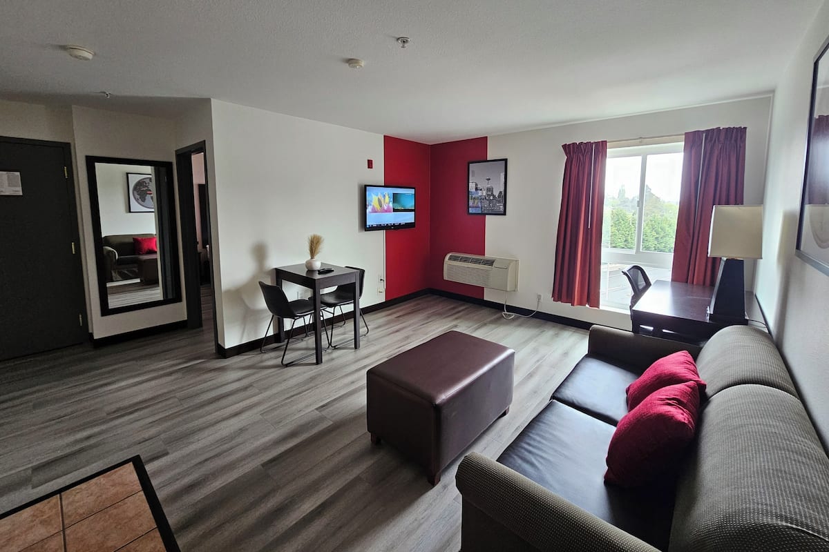 A modern hotel room with a dining area, a sofa, a TV, and a window with curtains.