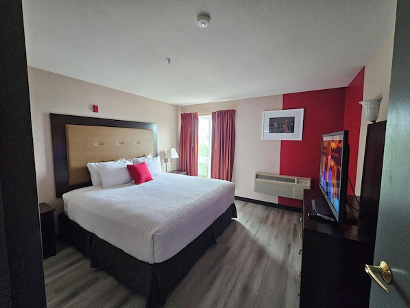 A hotel room with a large bed, a bedside table with a lamp, a window with red curtains, a framed picture on the wall, and a television.