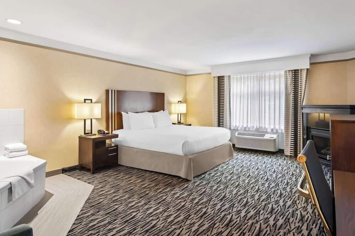 A hotel room with a bed, bedside lamps, a TV, a window with curtains, and a radiator.