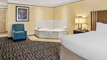 A hotel room with a large bed, a sitting area with a chair, a bathtub, a bedside table with a lamp, and a framed picture on the wall.