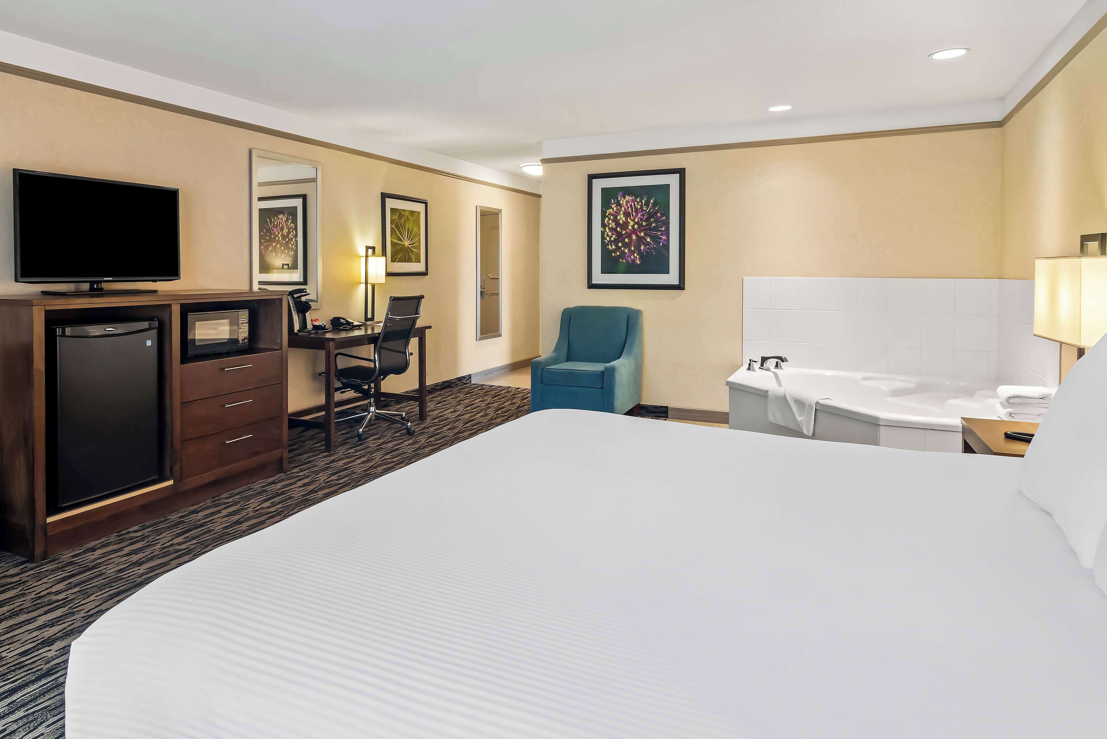 A hotel room with a large bed, a desk with a chair, a television, a microwave, a refrigerator, a bathtub, and a large framed picture on the wall.