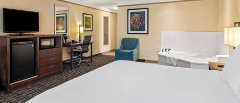 A hotel room with a large bed, a desk with a chair, a television, a microwave, a refrigerator, a bathtub, and a large framed picture on the wall.