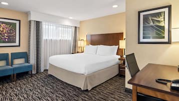 A hotel room with a large bed, a desk with a telephone, two blue armchairs, a wall-mounted TV, and a framed picture on the wall.