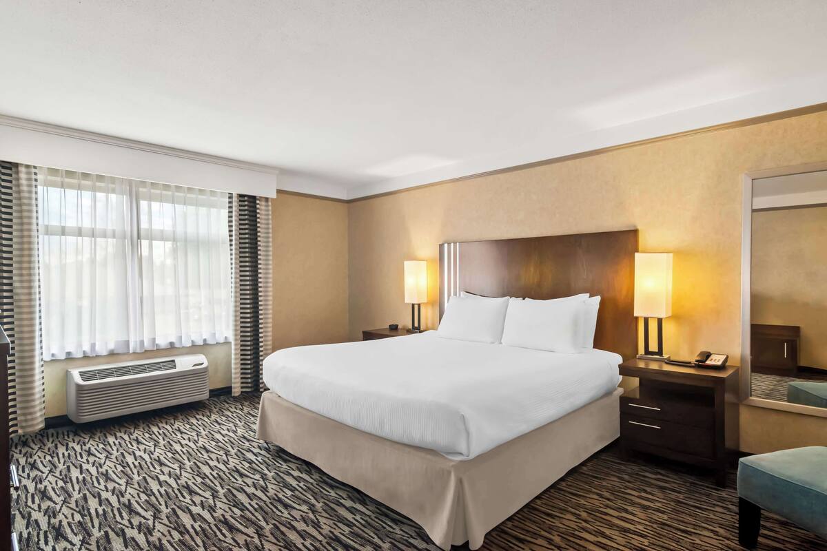 A hotel room with a large bed, two bedside lamps, a nightstand, a mirror, and a window with blinds.