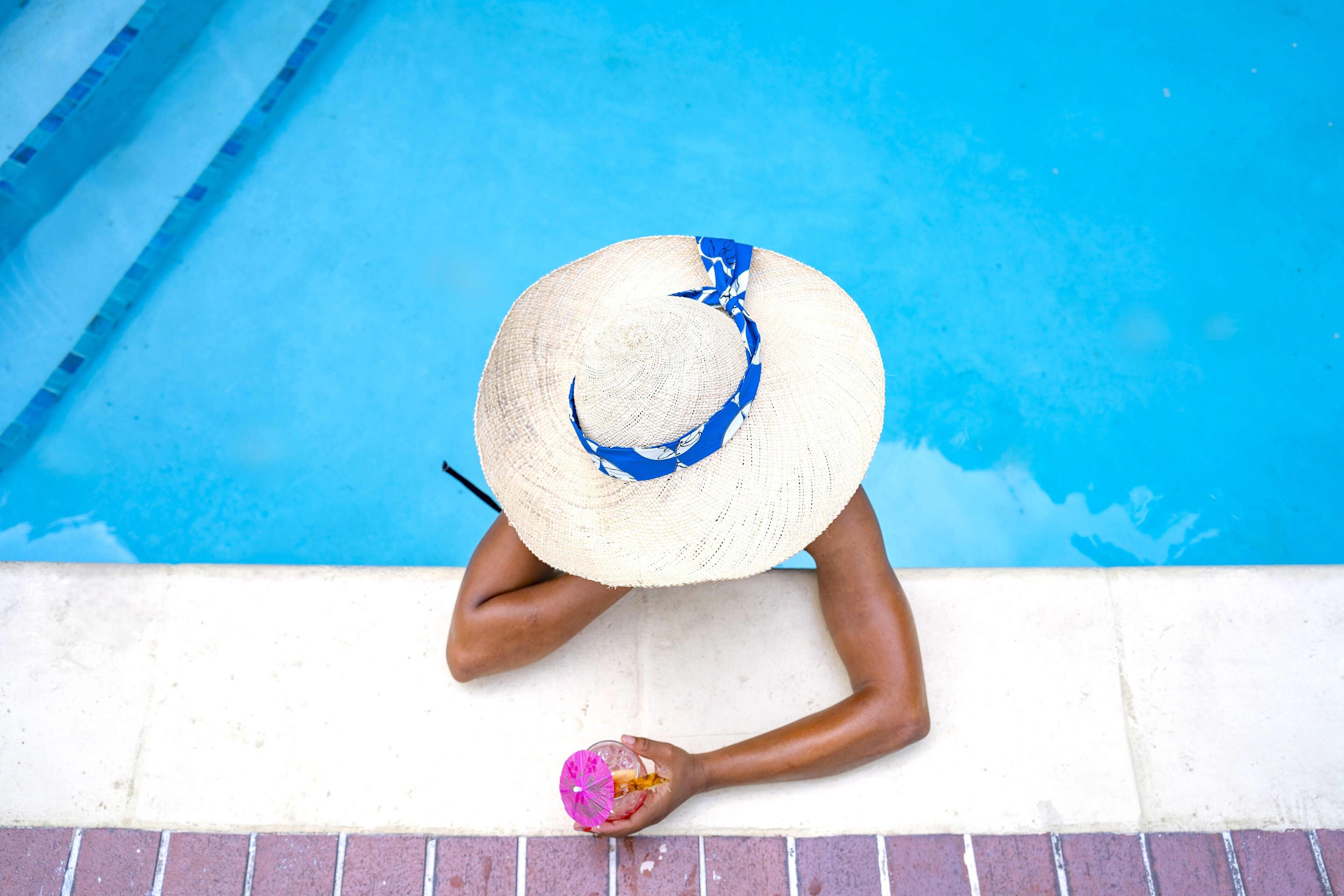 Eine Person sitzt am Pool, ein Strohhut und ein Getränk sind vorhanden.