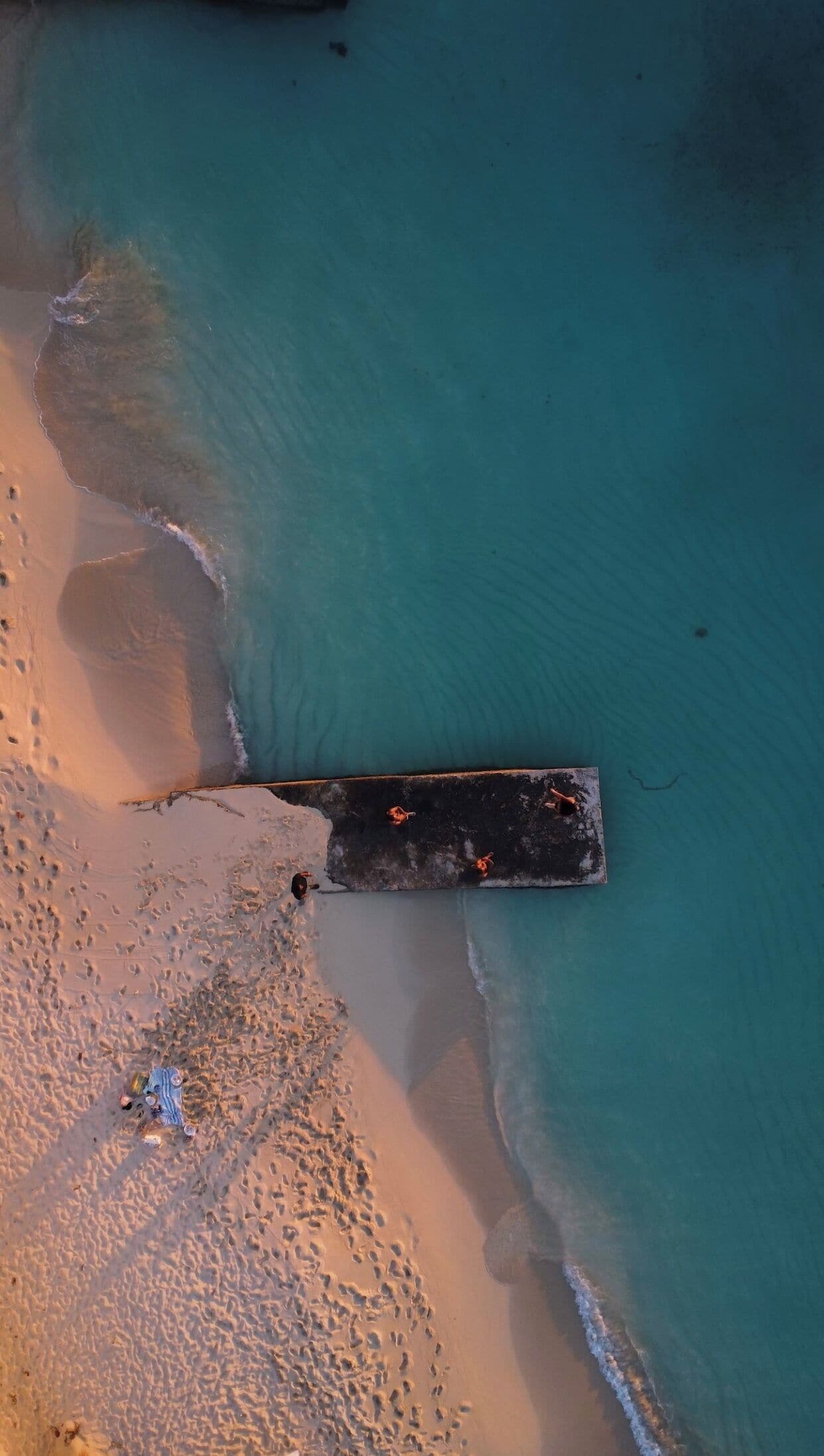 An aerial view of a beach with a wooden pier extending into the water, surrounded by footprints and a few scattered people.