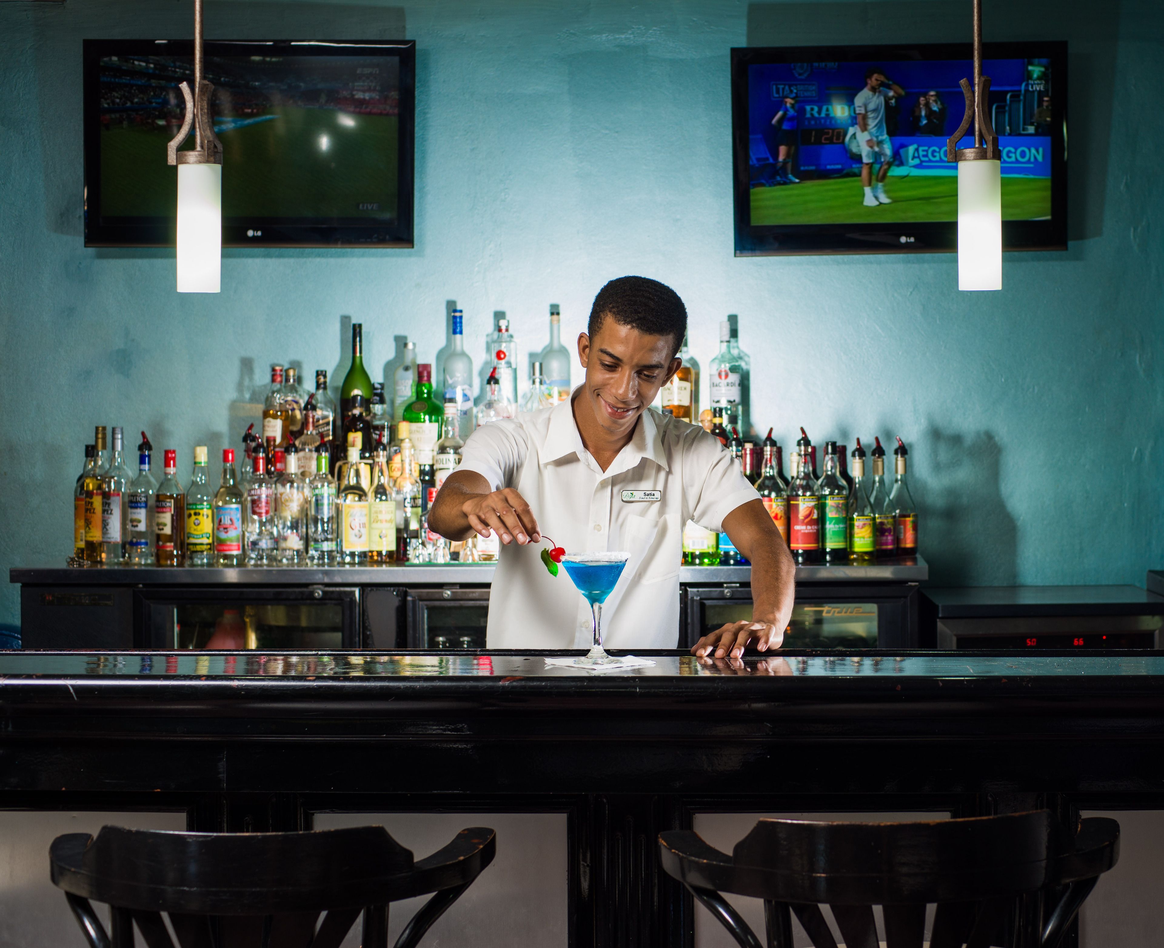 Un bar où un barman prépare un cocktail, des étagères remplies de bouteilles d’alcool et deux téléviseurs accrochés au mur.