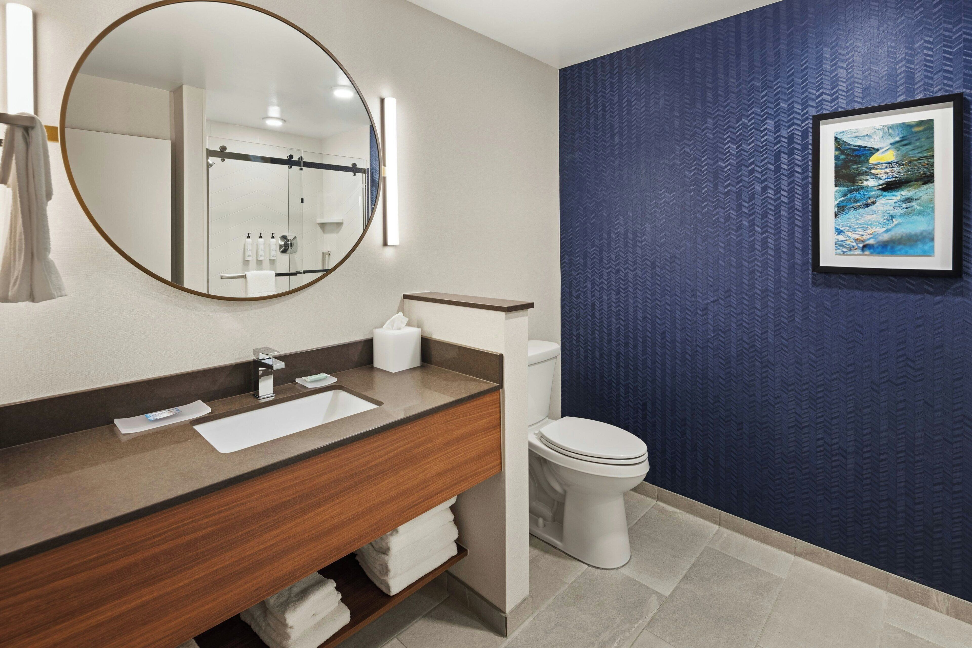 A modern bathroom with a wooden vanity, a round mirror, a toilet, and a blue textured wall.