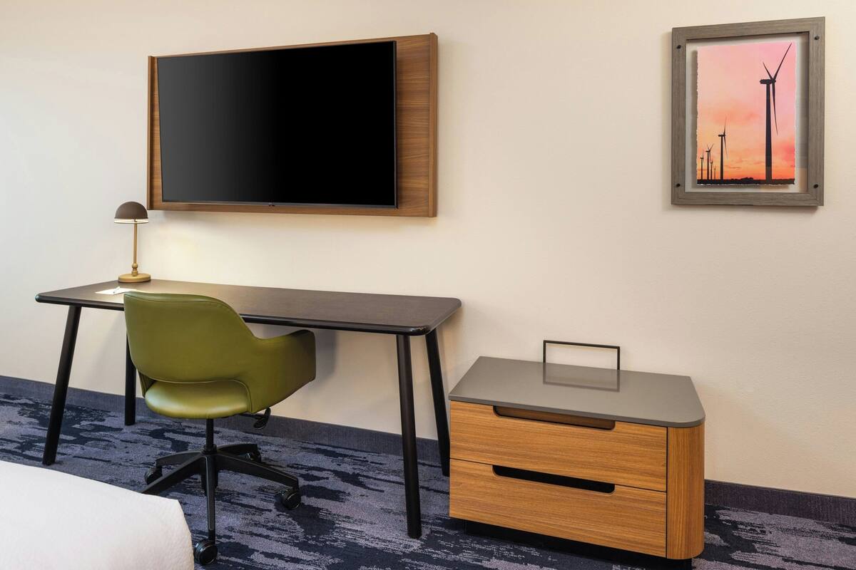 Une chambre d’hôtel équipée d’un téléviseur à écran plat fixé au mur, d’un bureau avec une chaise, d’une petite table d’appoint en bois et d’un tableau représentant des éoliennes.