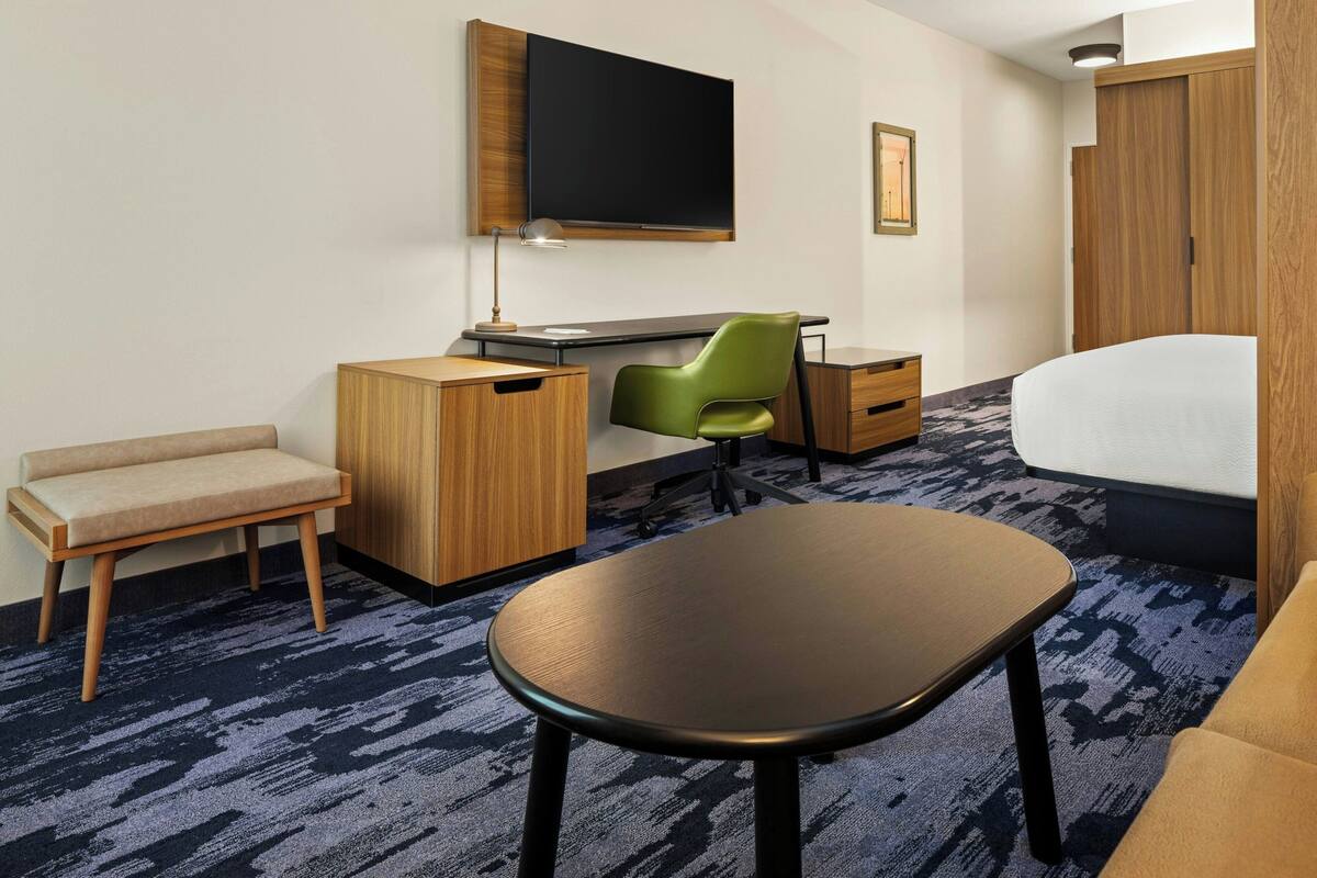 Une chambre d’hôtel avec un lit, un bureau avec un ordinateur, une chaise verte, un petit canapé et un téléviseur fixé au mur.