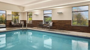 Piscina con bordes de azulejos, rodeada de sillas y mesas, ventanas amplias con vistas a árboles y una pared de acento de madera.
