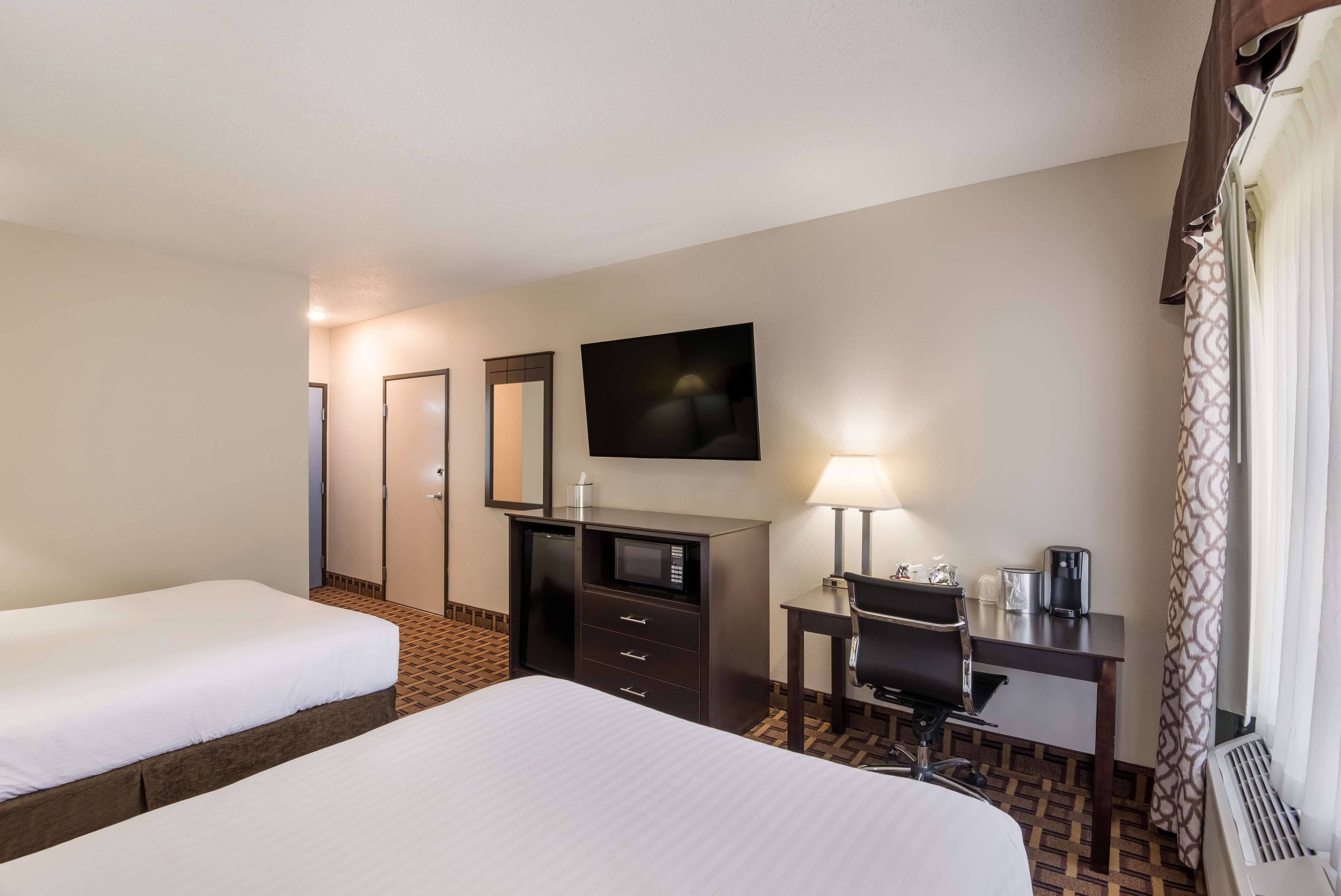 A hotel room with two beds, a desk with a chair, a microwave, a lamp, and a large mirror.