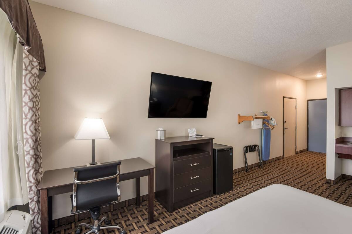 A hotel room with a bed, desk, chair, TV, lamp, and a small kitchenette.
