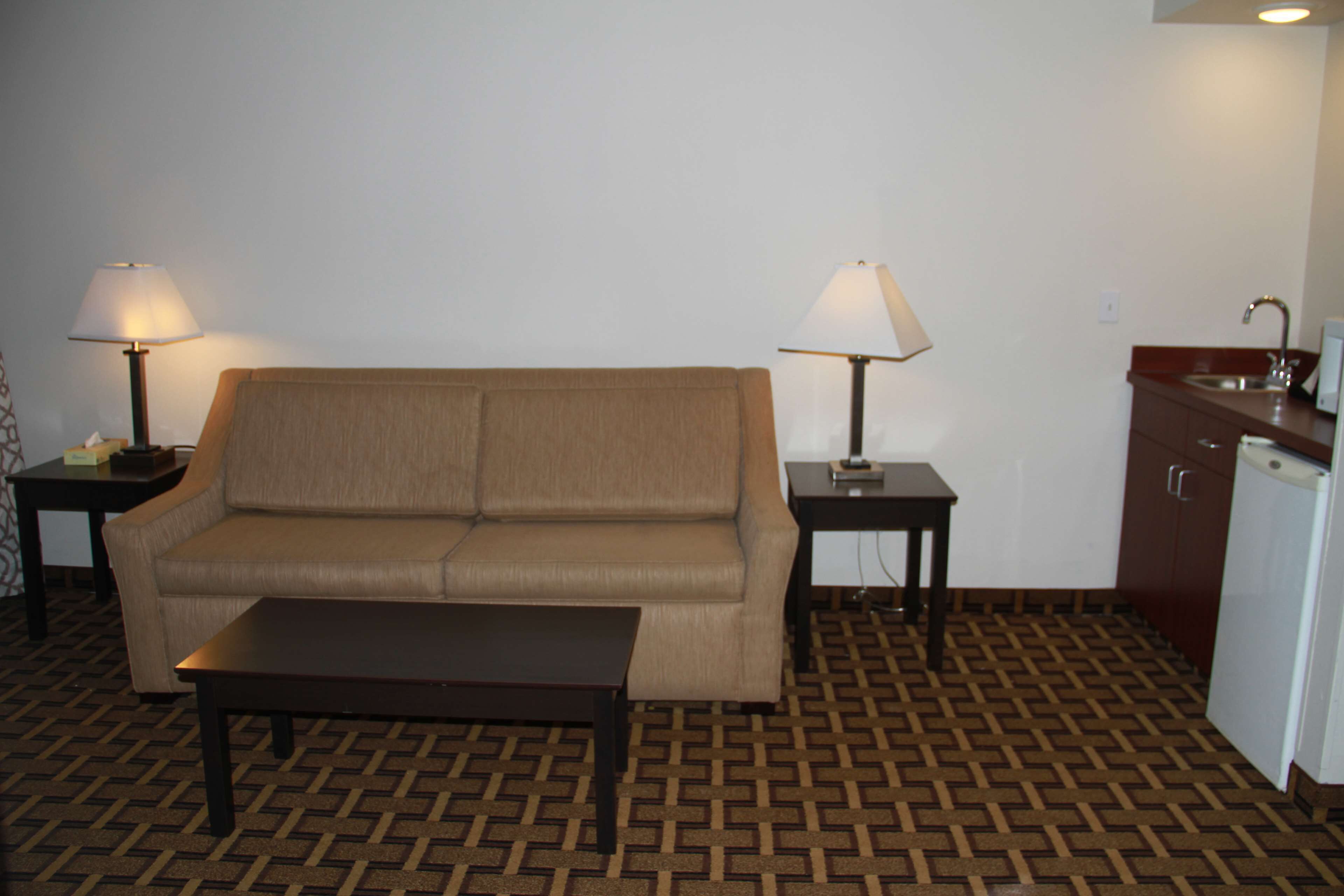 A small, furnished room with a sofa, a coffee table, two lamps, a small table, a sink, and a refrigerator.
