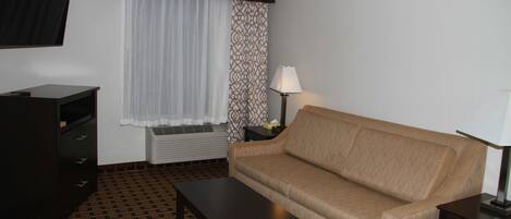 Ein Zimmer mit einem braunen Sofa, einem dunklen Holztisch, einem an der Wand montierten Fernseher und gemusterten Vorhängen.