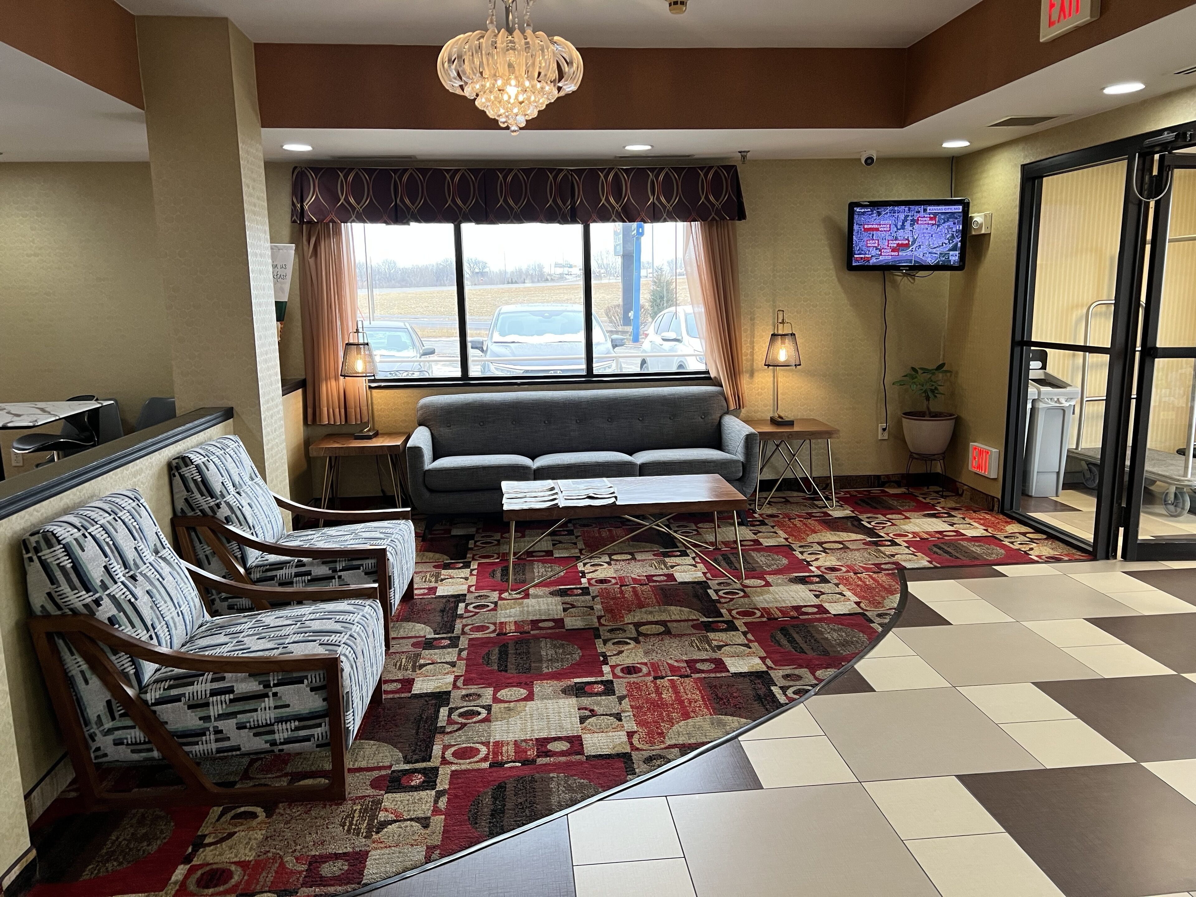 A hotel lobby with a sofa, chairs, a patterned carpet, a chandelier, a television, and a view of the parking lot.