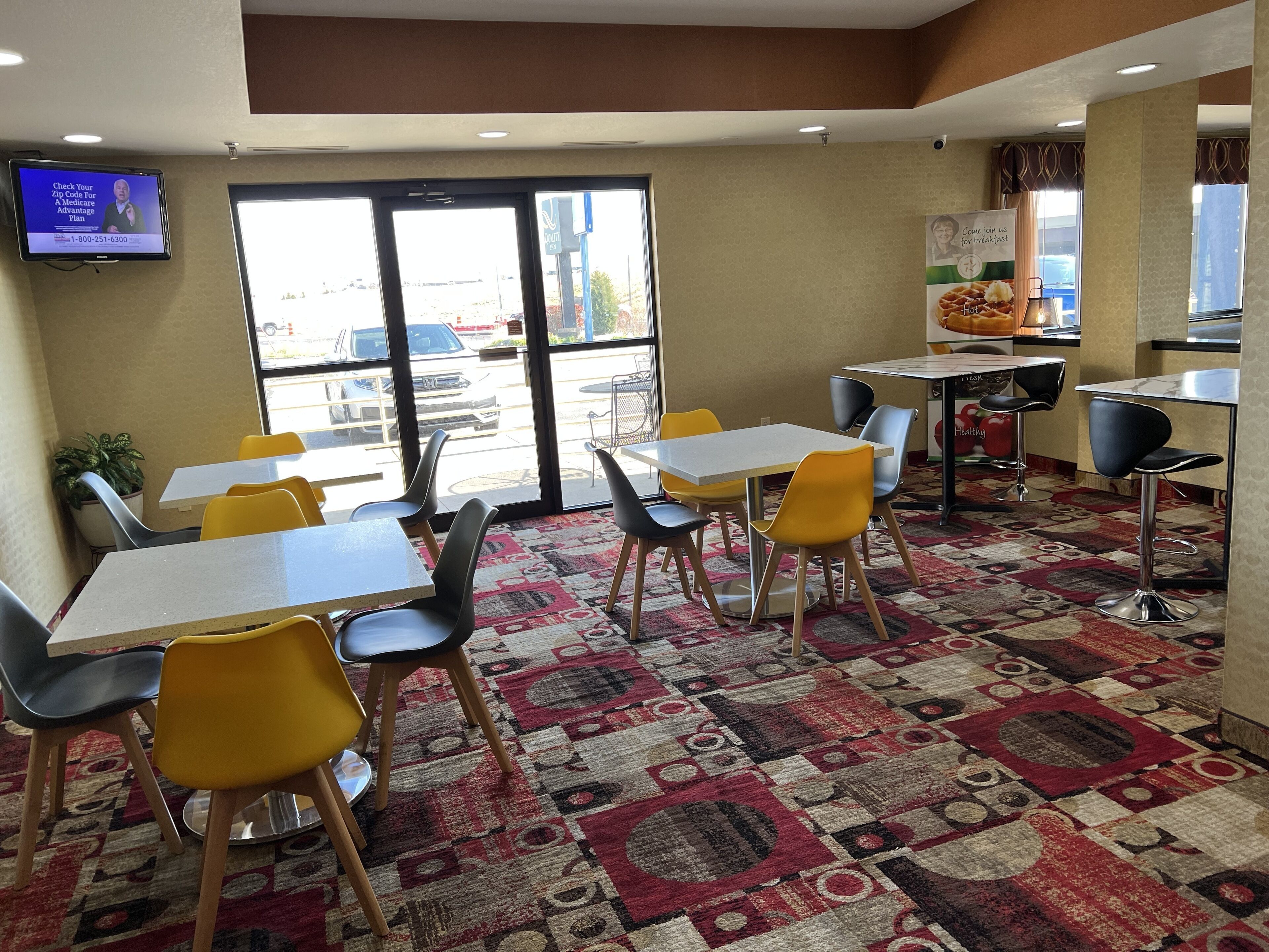 A restaurant with tables and chairs, a patterned carpet, and a television displaying a Medicare advertisement.
