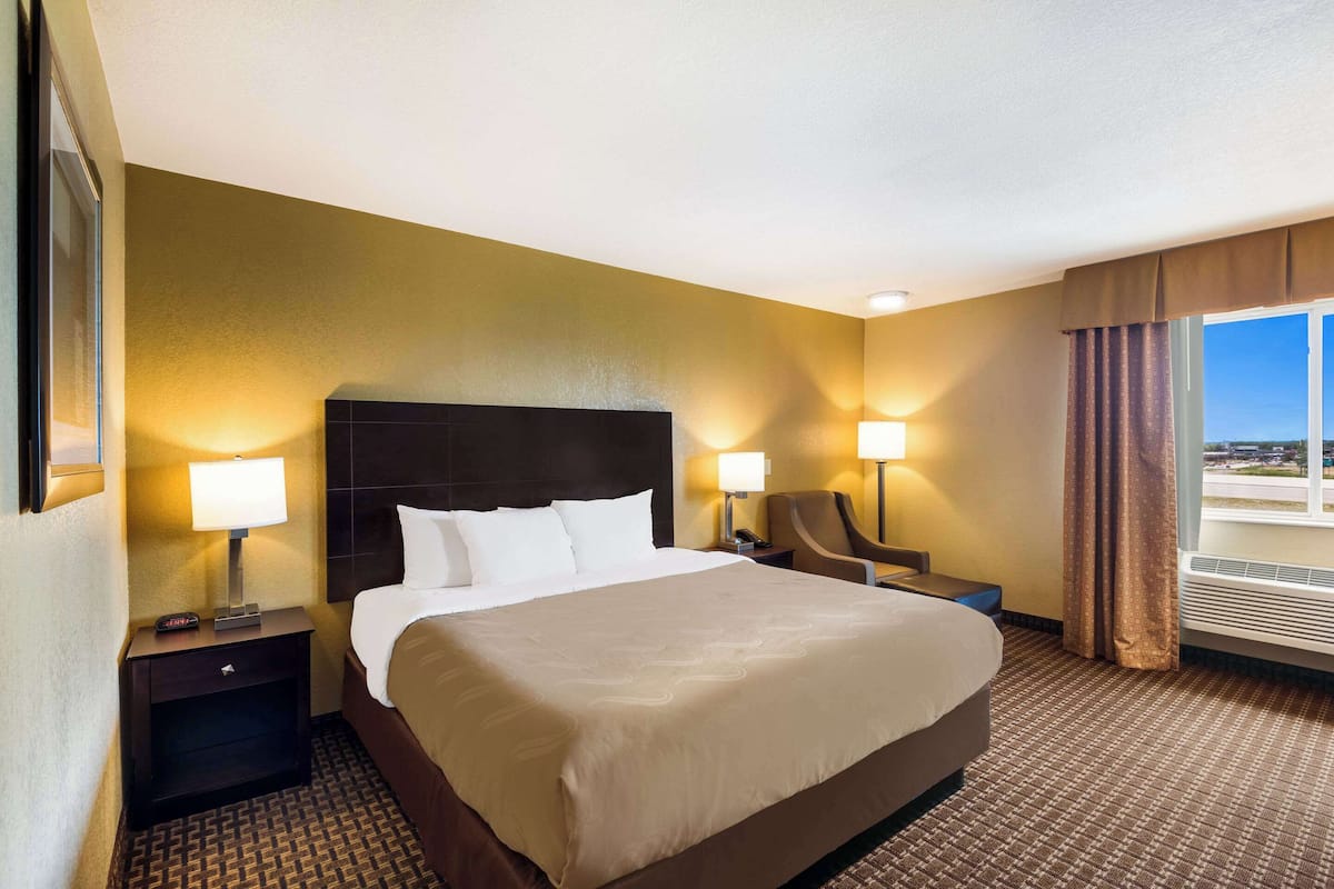 A hotel room with a large bed, two bedside lamps, a chair, a nightstand, and a window with curtains.