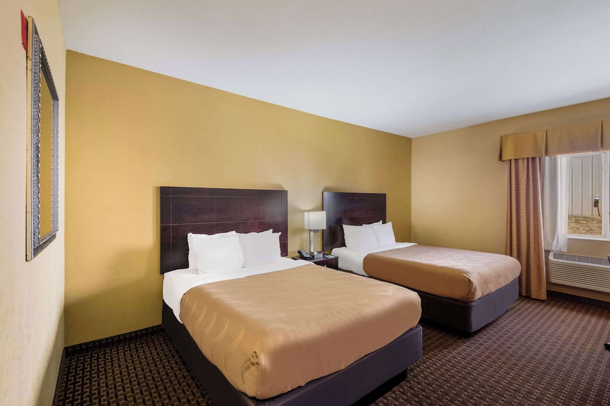 A hotel room with two beds, a nightstand, a lamp, and a window with curtains.