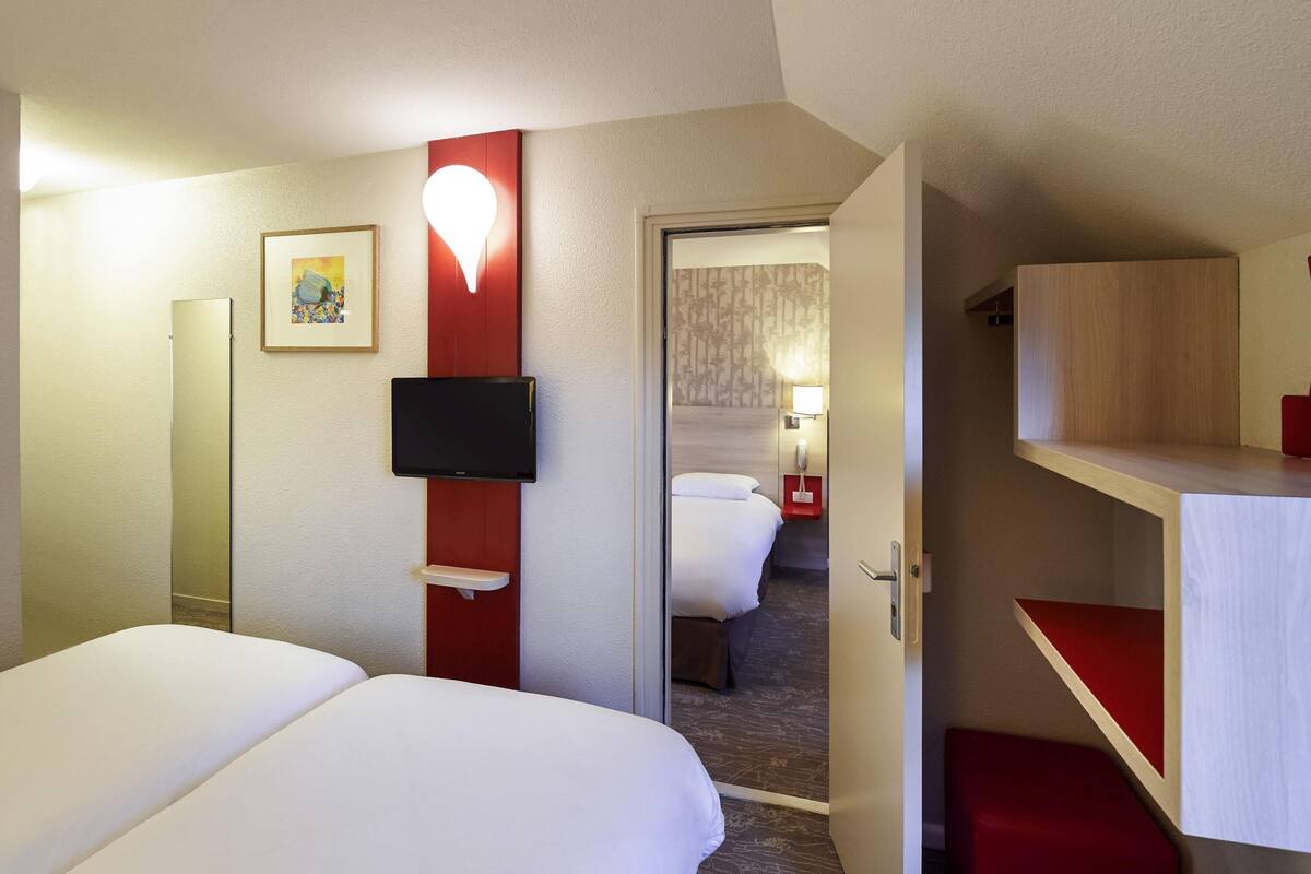 Hotelkamer met een bed, een aan de muur bevestigde televisie, een nachtkastje, een rode accentmuur en een deur naar een andere kamer.