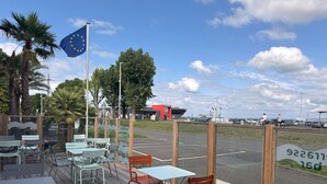 Een buitenzitplaats met tafels en stoelen, een vlag, en zicht op een parkeerplaats en gebouwen op de achtergrond.