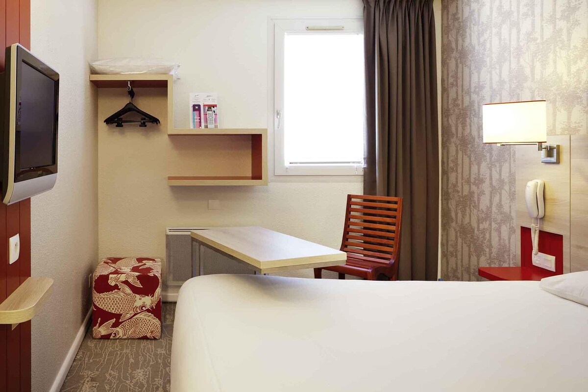 Habitación de hotel con cama, escritorio, silla, televisión, teléfono y ventana con cortinas.