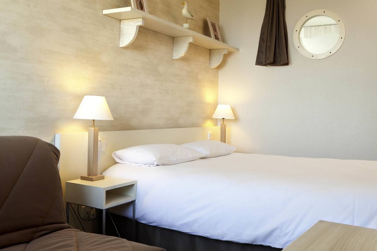 Une chambre d’hôtel comprenant un lit, deux lampes de chevet, une chaise, une petite table, un miroir rond et une étagère avec des objets décoratifs.