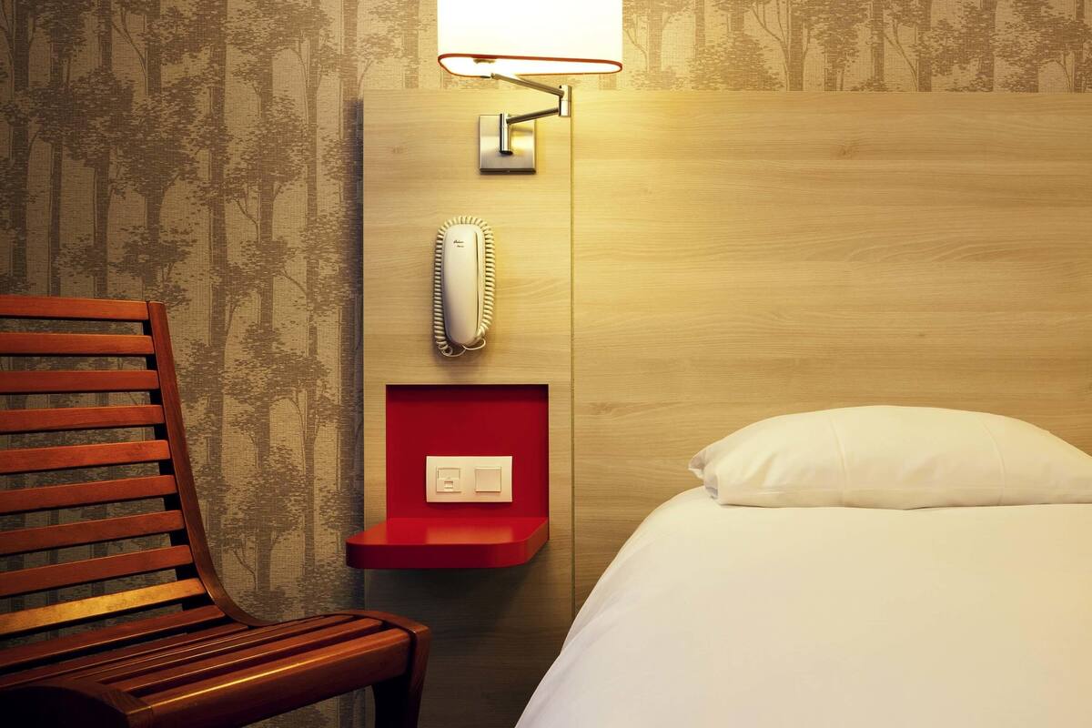 Habitación de hotel con cabecera de madera, una silla de madera, una mesita de noche con lámpara, teléfono y cama con ropa blanca.