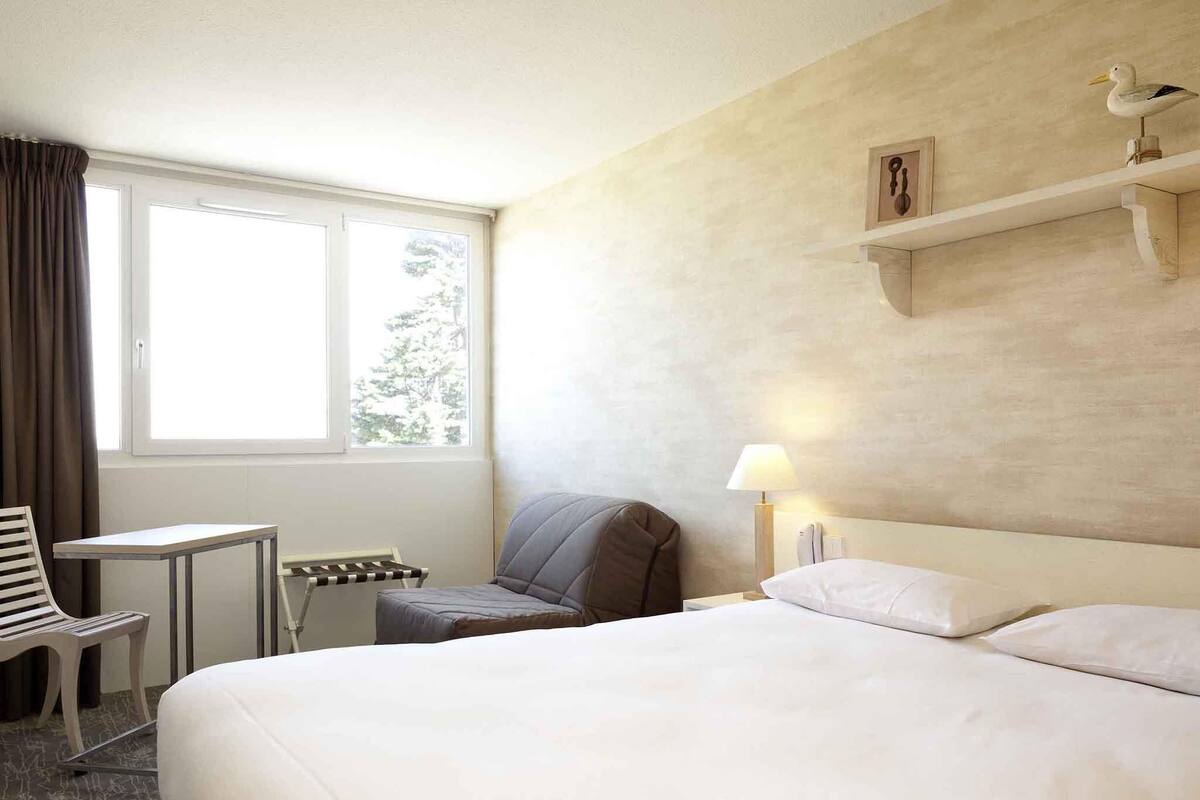 Een hotelkamer met een bed, een stoel, een tafeltje, een lamp en een raam met gordijnen.