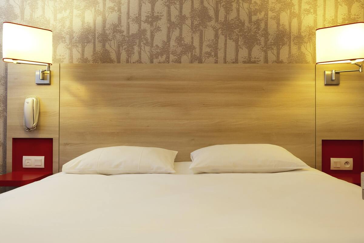 Une chambre d’hôtel avec une tête de lit en bois, deux lampes de chevet, un téléphone et un lit avec deux oreillers.