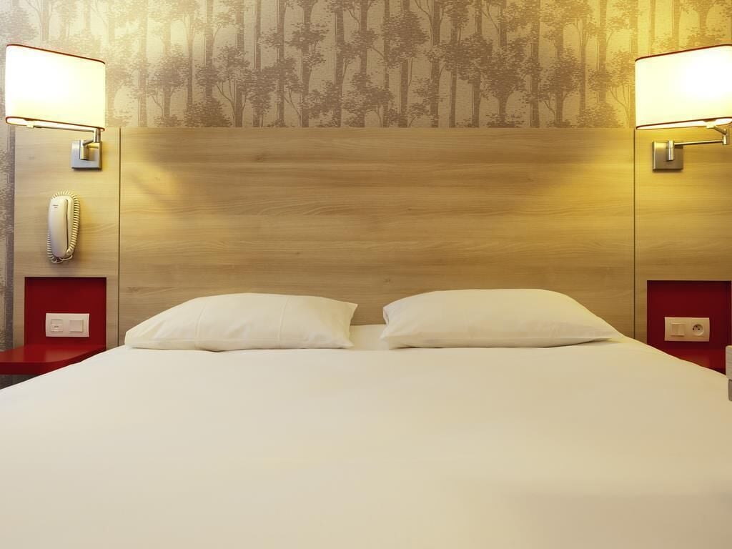 Habitación de hotel con cabecera de madera, dos almohadas blancas, una mesita de noche roja, un teléfono y una pared con diseño de árbol.