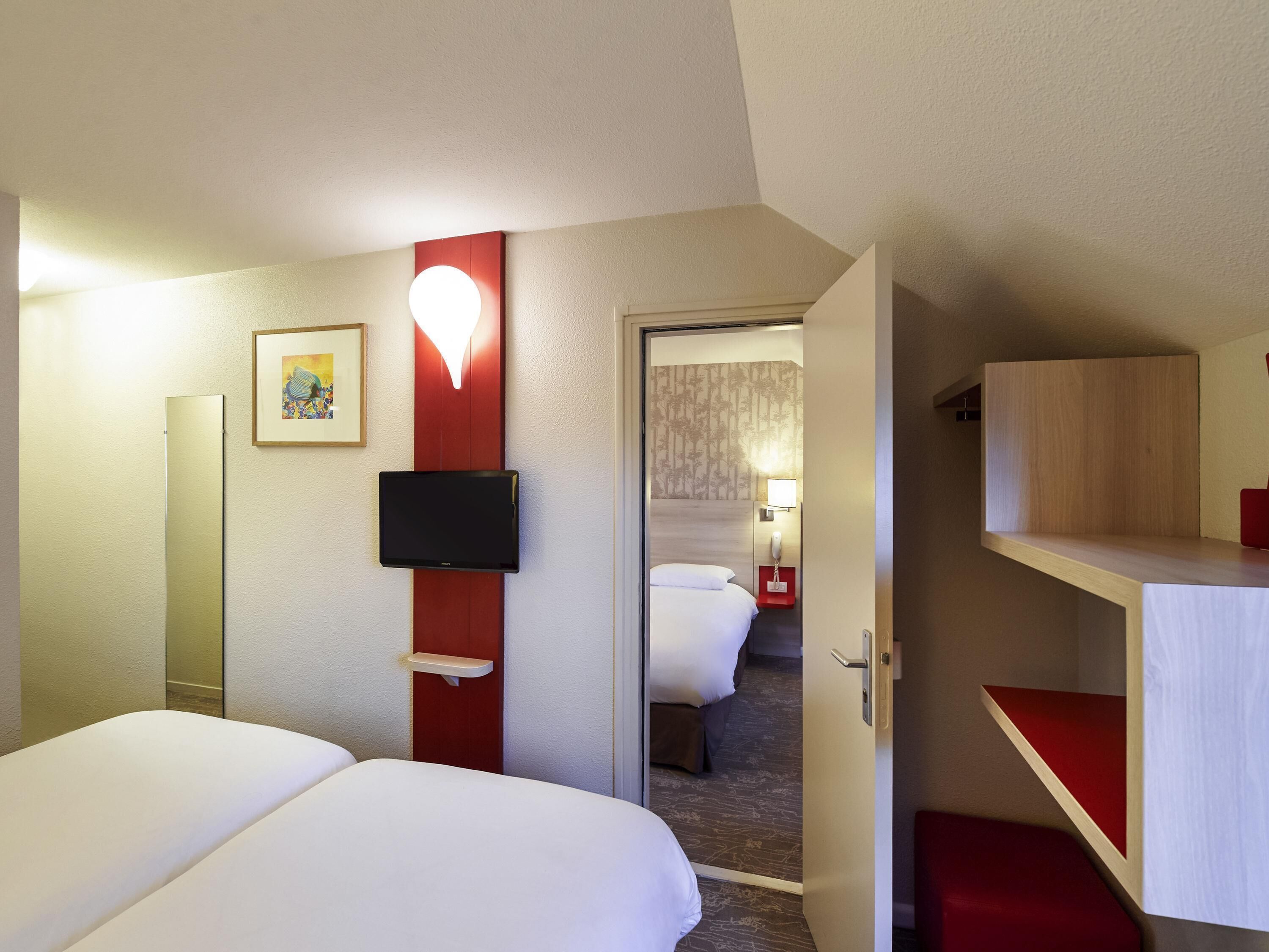 Hotelkamer met een bed, een aan de muur bevestigde televisie, een nachtkastje, een rode accentmuur en een deur naar een andere kamer.