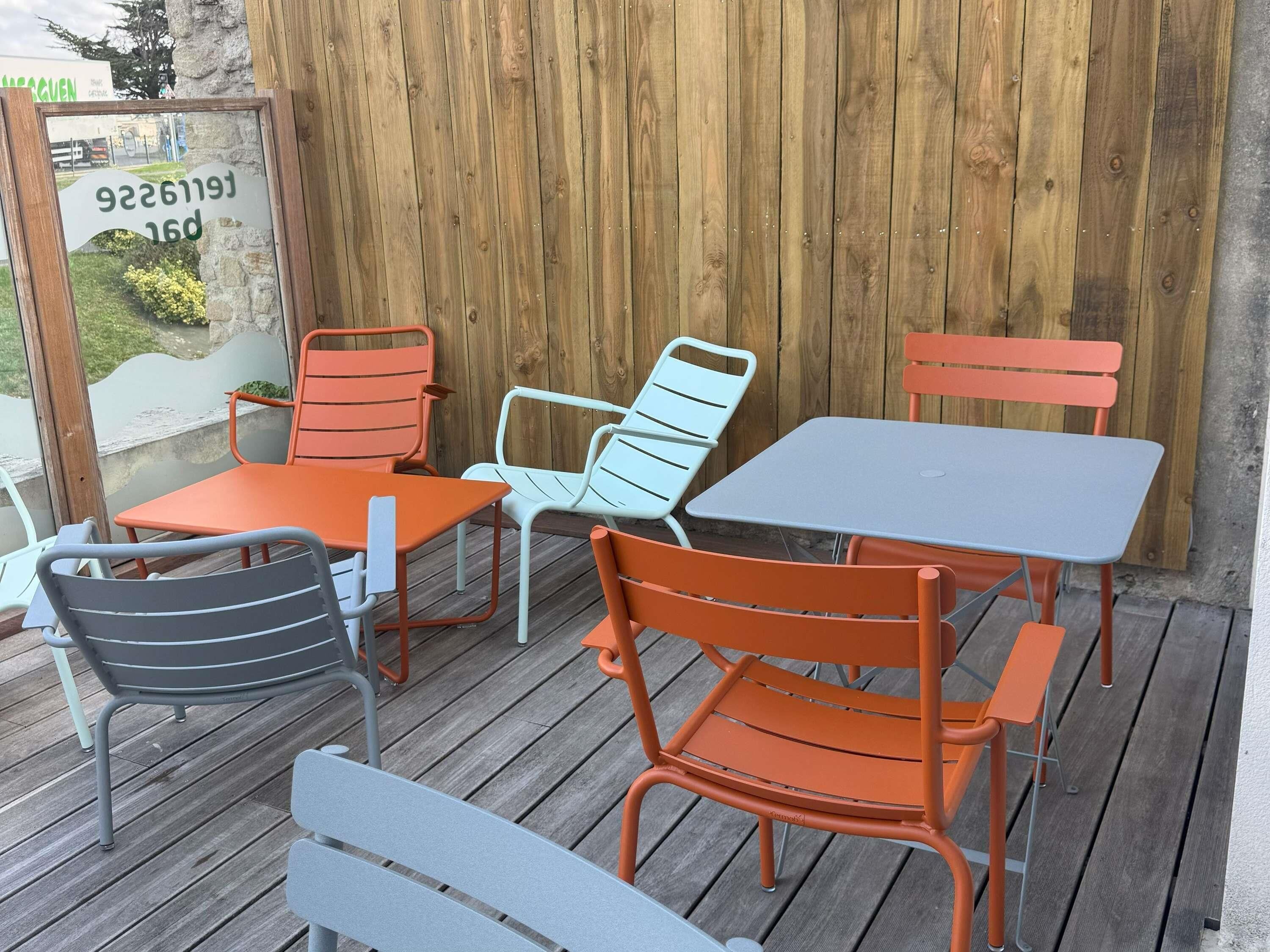 Una terraza de madera con sillas de metal y una mesa, un muro de piedra y un cartel con texto.