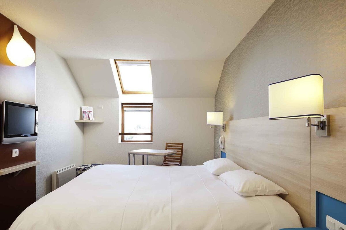 Una habitación de hotel con cama, escritorio, silla, televisor y ventana.