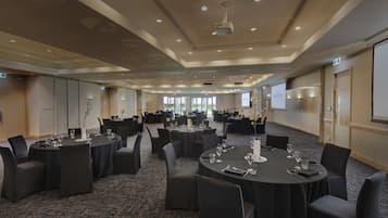 A spacious conference room with multiple round tables, each set with chairs and glassware, in a modern hotel setting.
