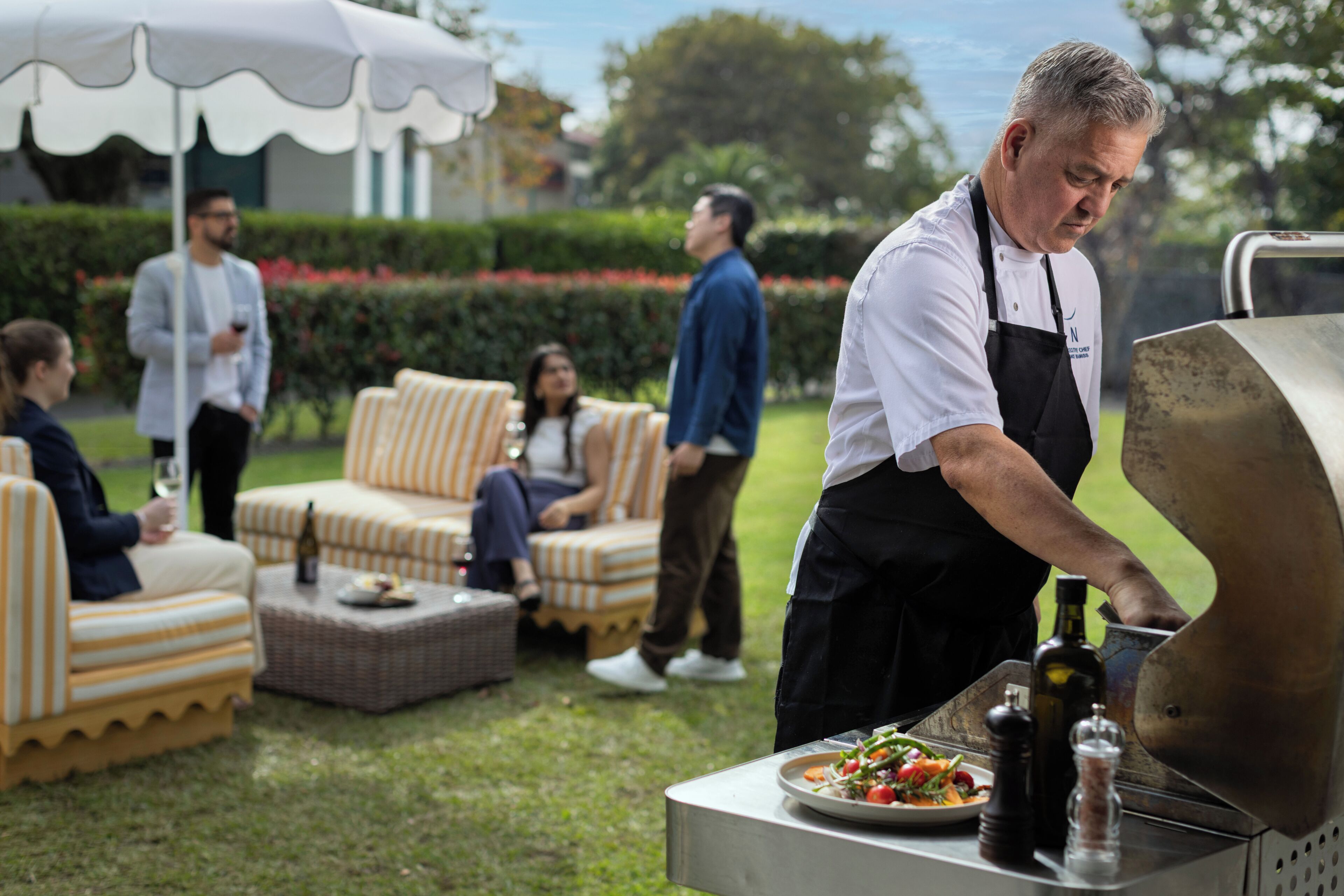 A chef grilling outdoors with a dining area in the background.