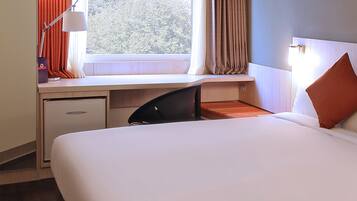 A hotel room with a bed, a desk with a chair, a lamp, and a window with curtains.