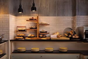 Un bufé de desayuno con panes y pasteles variados, una máquina de café y una pila de platos.